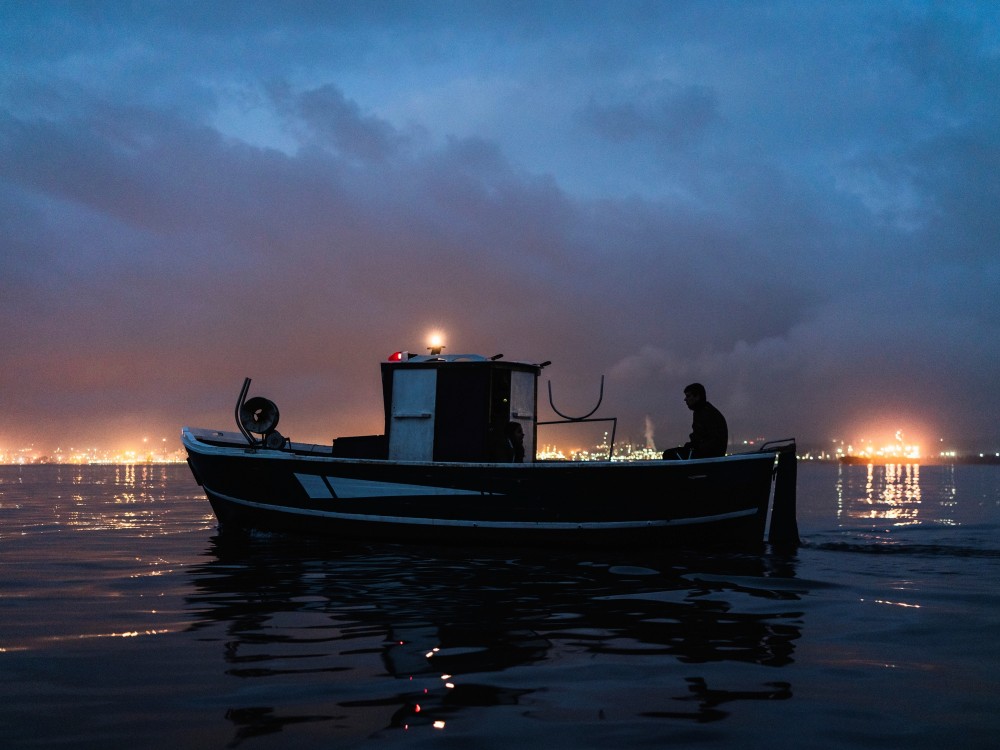 Ein Fischer fährt zu seinem Netz, während am Horizont die Raffinerien leuchten, Augusta, 2019